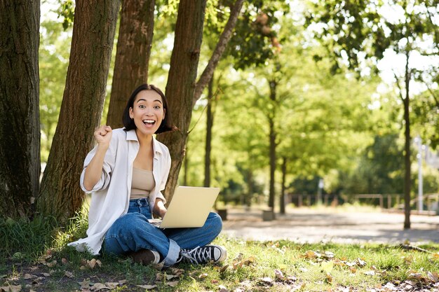 Retrato de una joven estudiante asiática haciendo deberes trabajando en un parque sentado al lado de un árbol con una laptop