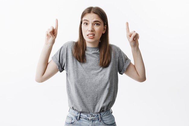 Retrato de joven estudiante atractiva morena en traje de moda haciendo expresión de la cara divertida, apuntando al revés con los dedos índices. Copie el espacio para la publicidad.