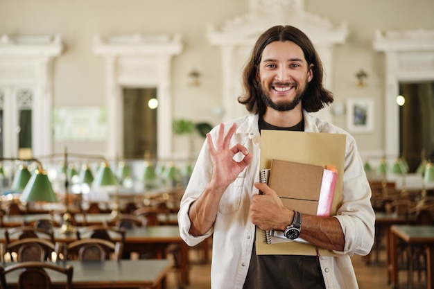 Retrato de un joven estudiante casual sonriente con un libro que muestra felizmente un gesto correcto en la biblioteca de la universidad