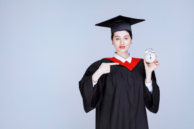 Retrato de joven estudiante graduada sosteniendo el reloj para mostrar el tiempo. Foto de alta calidad