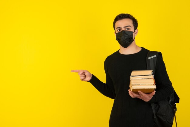 Retrato de joven estudiante en máscara médica sosteniendo libros universitarios y apuntando