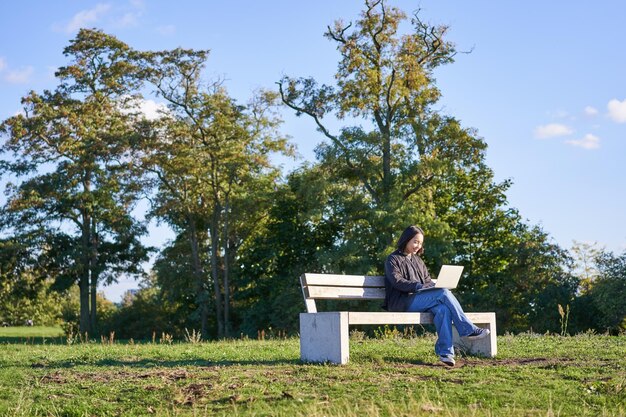 Retrato de una joven estudiante usando una laptop sentada en el parque en un banco escribiendo en la computadora