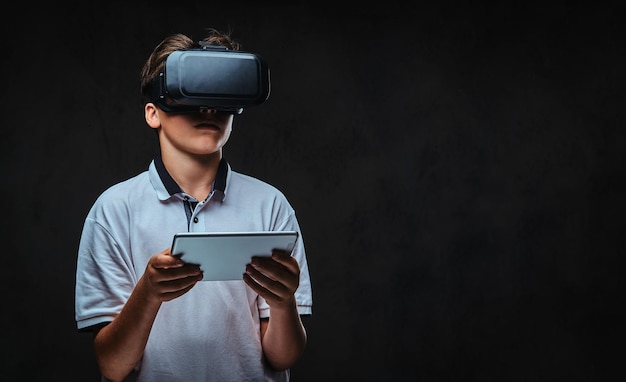 Retrato de un joven estudiante vestido con una camiseta blanca usando gafas de realidad virtual y una tableta. Aislado en un fondo oscuro.