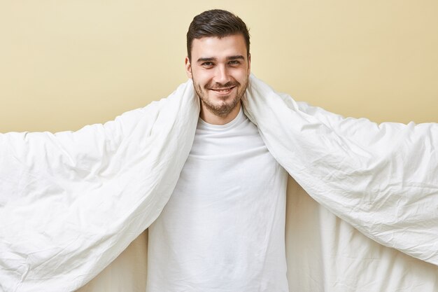 Retrato de joven europeo alegre y guapo huy con amplia sonrisa radiante sosteniendo una gran manta blanca con los brazos extendidos, y finalmente se va a dormir después de un duro día de trabajo