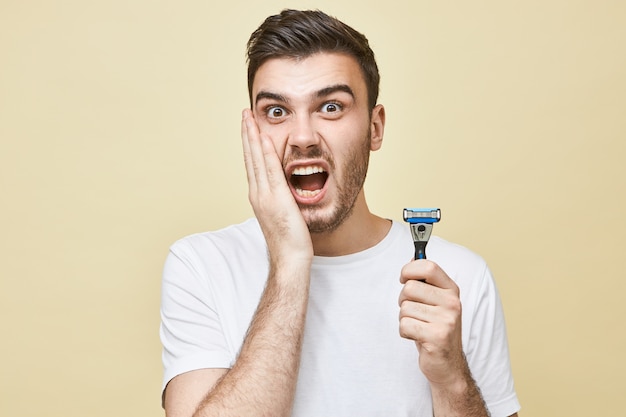 Retrato de un joven europeo frustrado e infeliz con barba que sostiene la mano en la mejilla y gritando, con mirada aterrorizada, que sufre de irritación de la piel debido al afeitado, sosteniendo una maquinilla de afeitar