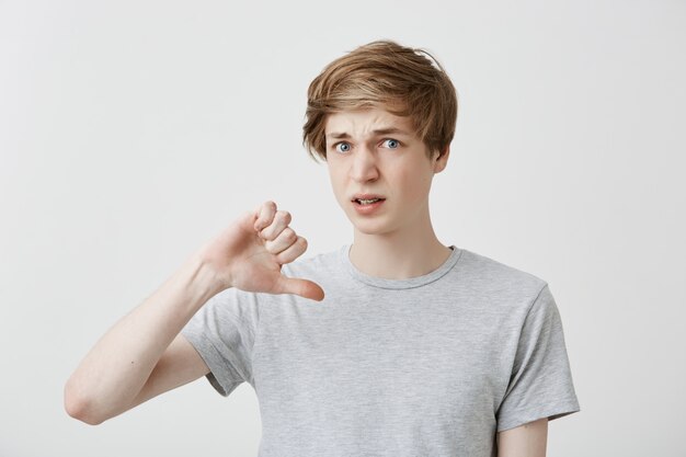 Retrato de joven europeo mantiene el pulgar hacia abajo, desaprueba algo, frunce el ceño con descontento. El chico rubio guapo luce una camiseta gris que no le gusta la película y no recomienda verla