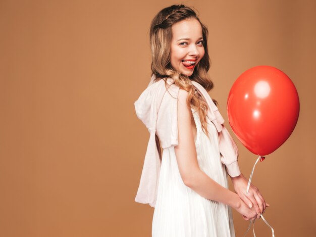 Retrato de joven excitada posando en vestido de verano blanco de moda. Mujer sonriente con globo rojo posando. Modelo lista para la fiesta, mostrando su lengua