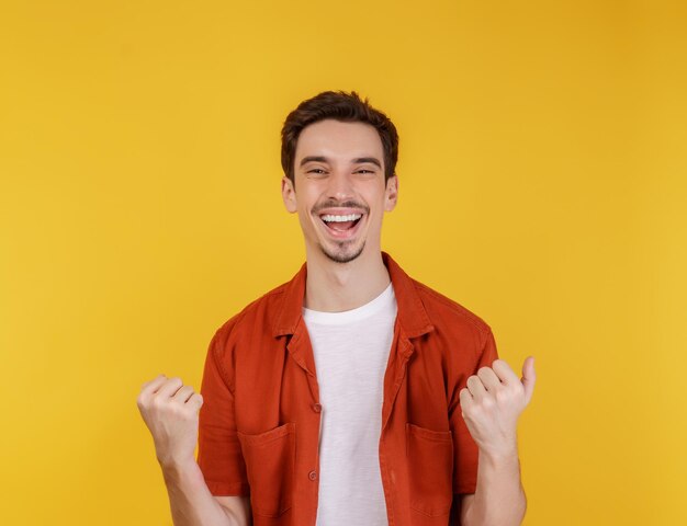 Retrato de un joven feliz y alegre de pie haciendo un gesto de ganador apretando los puños manteniéndose aislado en un estudio de fondo de pared de color amarillo