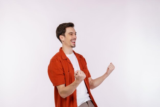 Retrato de un joven feliz y alegre de pie haciendo un gesto de ganador apretando los puños manteniéndose aislado en un estudio de fondo de pared de color blanco