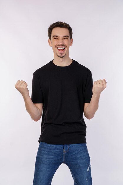 Retrato de un joven feliz y alegre de pie haciendo un gesto de ganador apretando los puños manteniéndose aislado en un estudio de fondo de pared de color blanco