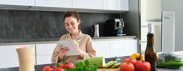 Foto gratuita retrato de una joven feliz escribe el menú para la cena se sienta en la cocina cerca de las verduras