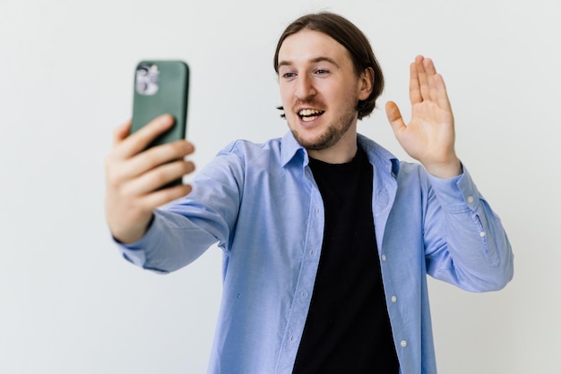 Retrato de un joven feliz gesticulando con el signo de la paz mientras usa un celular aislado sobre fondo blanco