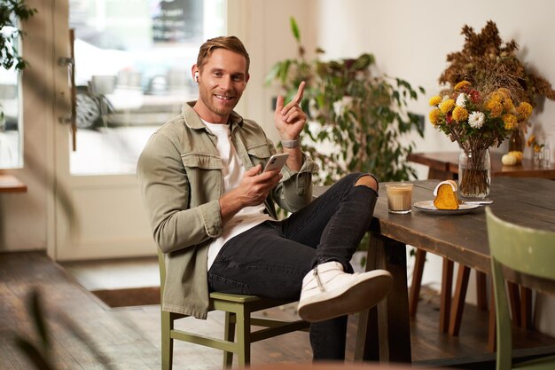 Retrato de un joven feliz y guapo sentado en un café con un teléfono móvil y auriculares inalámbricos