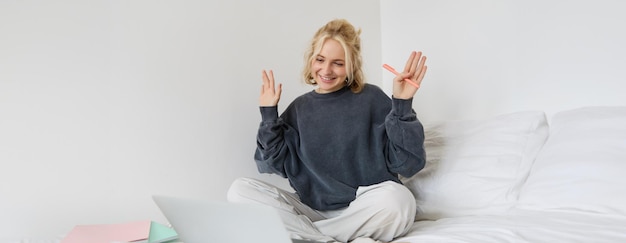 Foto gratuita retrato de una joven feliz que se conecta a un chat de video usando una computadora portátil agitando la mano a la cámara diciendo hola