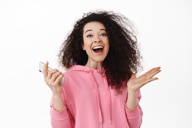 Retrato de una joven feliz que usa un teléfono inteligente, se regocija y grita de alegría, gana dinero en el teléfono móvil, triunfa, gana en el teléfono inteligente, se enfrenta a un fondo blanco