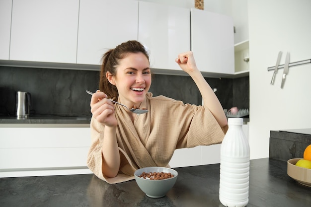 Retrato de una joven feliz riendo comiendo cereales con leche triunfando desayunando y