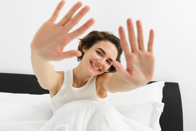 Retrato de una joven feliz y soñolienta que se despierta en su cama extendiendo las manos escondiendo la cara de