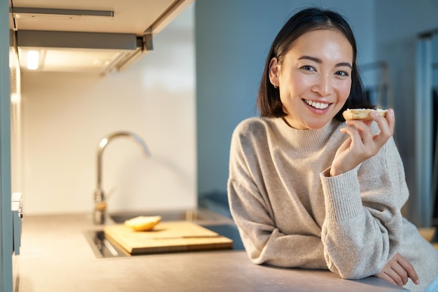 Retrato de una joven feliz sonriente que se queda en casa parada en la cocina y comiendo tostadas mirando a