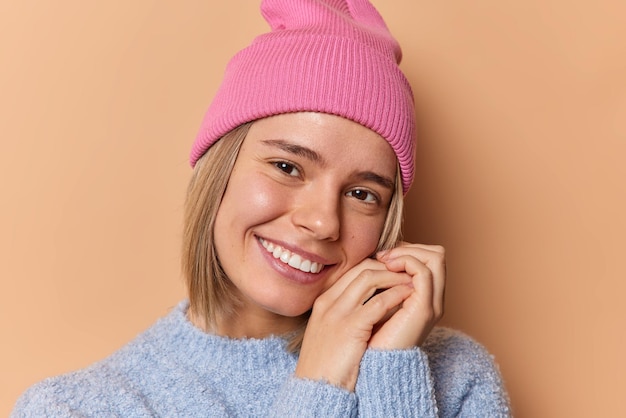 Retrato de una joven feliz y tierna inclina la cabeza y sonríe con dientes mantiene las manos cerca de la cara usa sombrero y un jersey casual aislado sobre fondo beige admira algo Expresiones faciales agradables