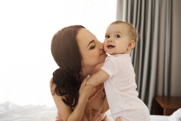 Retrato de joven feliz tierna madre y su pequeña hija sonriente sentada en la cama en su casa