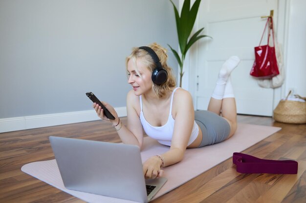 Retrato de una joven de fitness que elige hacer ejercicio en una computadora portátil con auriculares y usando un teléfono móvil