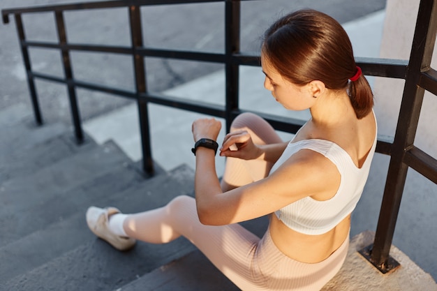 Foto gratuita retrato de una joven fitness sentada en las escaleras al aire libre y comprobando la cantidad de calorías quemadas después del entrenamiento, mujer de pelo oscuro en top blanco y leggins beige.