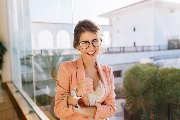 Retrato de joven con gafas elegantes, dama inteligente en elegante chaqueta rosa con blusa beige, estudiante feliz mostrando los pulgares para arriba. Ventana grande con linda vista.