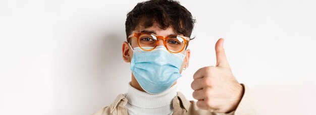 Retrato de un joven con gafas y máscara médica que muestra el pulgar hacia arriba sonriendo con los ojos parados en whi