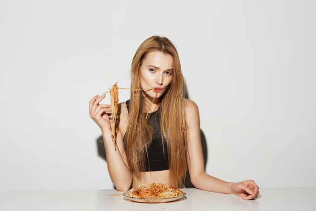 Retrato de una joven guapa con el pelo largo y claro en la parte superior negra comiendo espaguetis con un tenedor mirando a la cámara con una mirada relajada