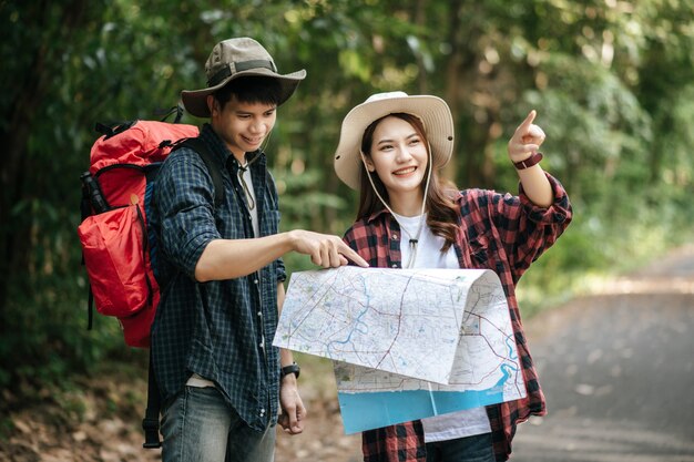 Retrato joven guapo asiático con mochila y sombrero de trekking y novia bonita de pie y comprobando la dirección en el mapa de papel mientras camina por el sendero del bosque, concepto de viaje con mochila