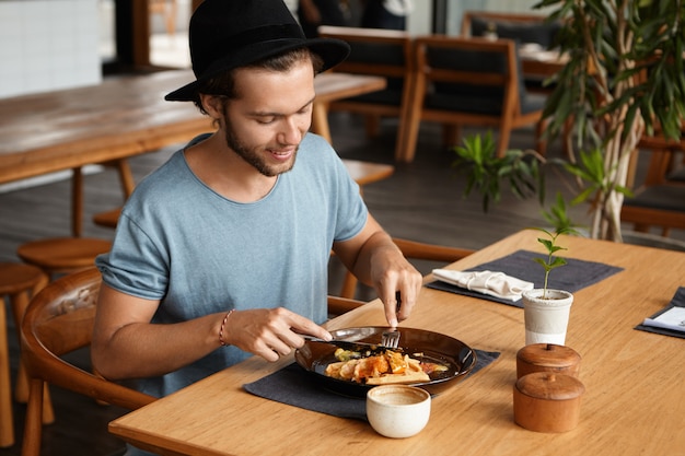 Retrato de joven guapo con barba sonriendo felizmente mientras come una sabrosa comida con cuchillo y tenedor