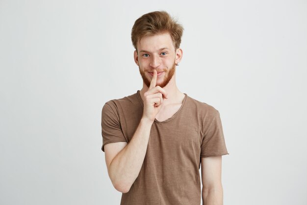 Retrato de joven guapo con barba sonriendo mostrando para guardar silencio.