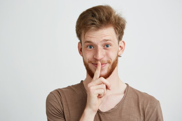 Retrato de joven guapo con barba sonriendo mostrando para guardar silencio.