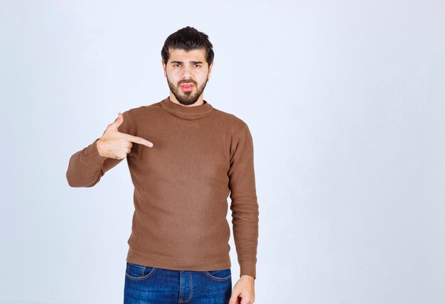 Retrato de joven guapo con barba en suéter marrón de pie y apuntando a sí mismo. Foto de alta calidad