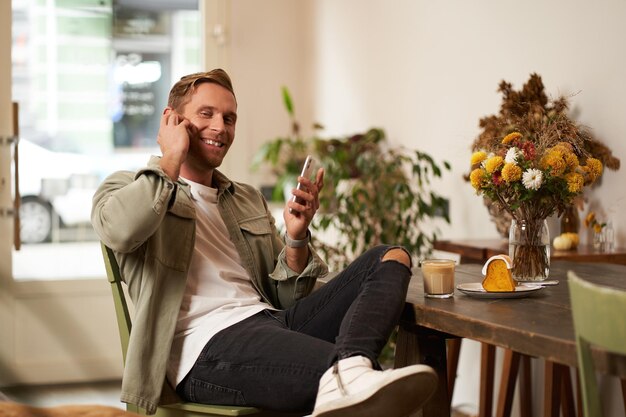 Retrato de un joven guapo y elegante visitante de un café sentado con su teléfono inteligente y escuchando