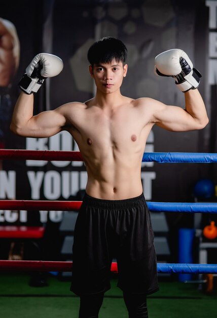 Retrato de joven guapo en guantes de boxeo blancos pose de pie sobre lienzo en el gimnasio, levanta los brazos hacia arriba para mostrar el músculo perfecto, clase de boxeo de entrenamiento de hombre sano,