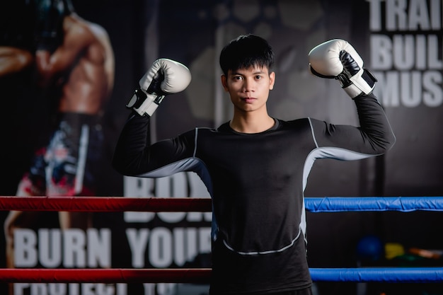 Retrato de joven guapo en ropa deportiva y guantes de boxeo blancos de pie levantar los brazos para posar sobre lienzo en el gimnasio, clase de boxeo de entrenamiento de hombre sano,