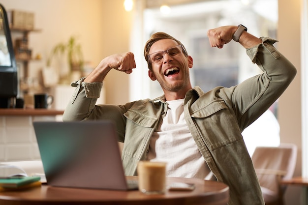 Foto gratuita retrato de un joven guapo y satisfecho que muestra los bíceps flexionando los músculos sentado frente a la computadora portátil en una cafetería