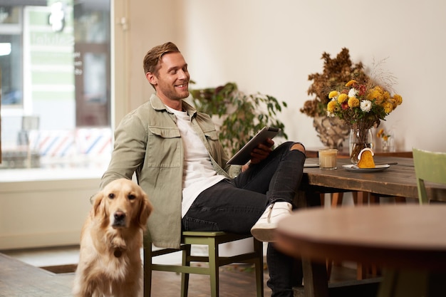 Foto gratuita retrato de un joven guapo sentado en un café con su perro acariciando a un golden retriever leyendo noticias en