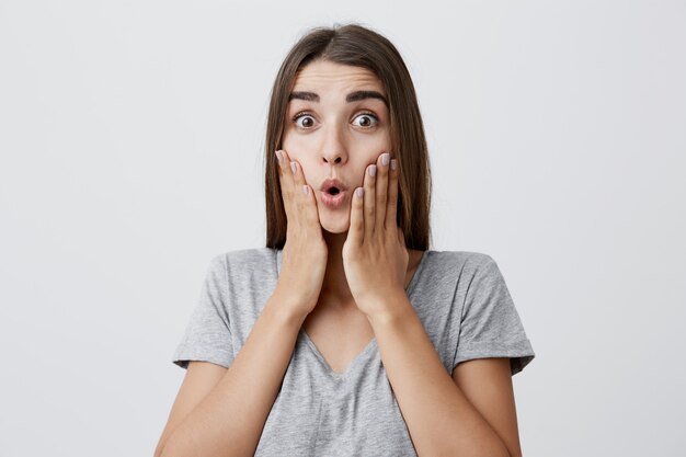 Retrato de joven hermosa caucásica estudiante divertida niña con cabello largo y castaño en traje de moda apretando la cara con las manos con la boca abierta y las cejas levantadas