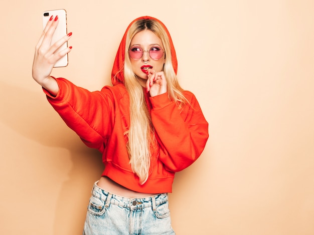 Retrato de joven hermosa chica mala inconformista en ropa de jeans de moda y aretes en la nariz. Mujer rubia sonriente despreocupada sexy toma selfie. Modelo positivo lamiendo dulces de azúcar redondo