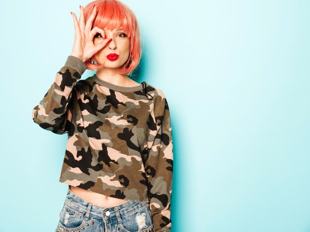 Retrato de joven hermosa chica mala inconformista en ropa de verano rojo de moda y arete en la nariz. Mujer sonriente despreocupada sexy posando en el estudio con peluca rosa. El modelo cubre su ojo y muestra un signo bien