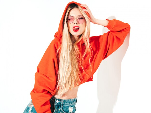 Retrato de joven hermosa chica mala inconformista en sudadera con capucha roja de moda y arete en la nariz. Modelo positivo divirtiéndose Muestra lengua