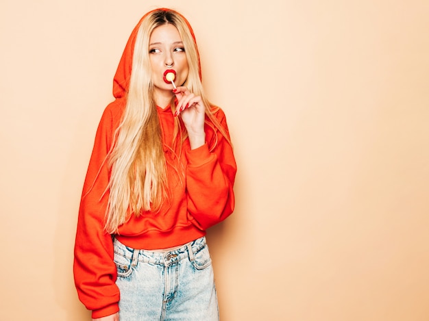 Retrato de joven hermosa chica mala inconformista en sudadera con capucha roja de moda y arete en la nariz. Modelo positivo lamiendo caramelos de azúcar redondos