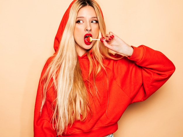 Foto gratuita retrato de joven hermosa chica mala inconformista en sudadera con capucha roja de moda y arete en la nariz. modelo positivo lamiendo caramelos de azúcar redondos