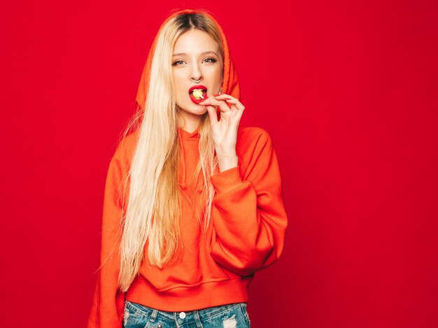 Retrato de joven hermosa chica mala inconformista en sudadera con capucha roja de moda y arete en la nariz. Modelo positivo lamiendo caramelos de azúcar redondos