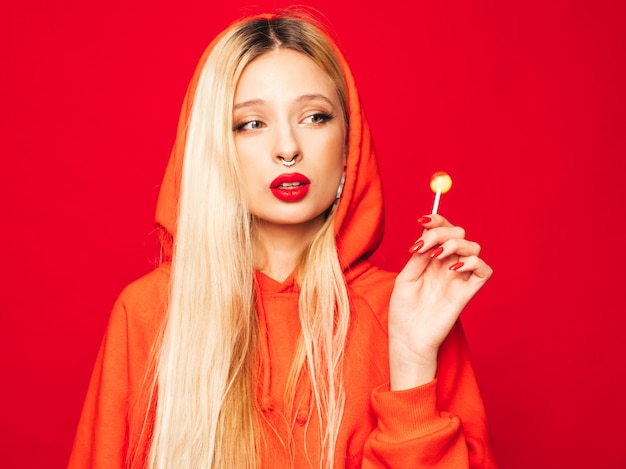 Retrato de joven hermosa chica mala inconformista en sudadera con capucha roja de moda y arete en la nariz. Modelo positivo lamiendo caramelos de azúcar redondos