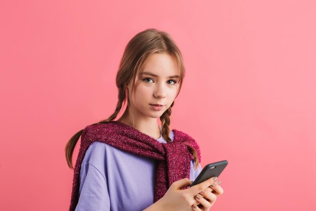 Retrato de una joven hermosa con dos trenzas en una camiseta lila y un suéter en los hombros sosteniendo un celular en la mano mirando cuidadosamente a la cámara sobre un fondo rosado aislado