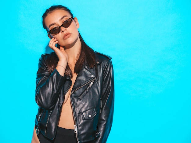 Retrato de joven hermosa hipster chica mala en moda verano negro chaqueta de cuero y gafas de sol. Mujer despreocupada atractiva aislada en azul. Modelo morena posando