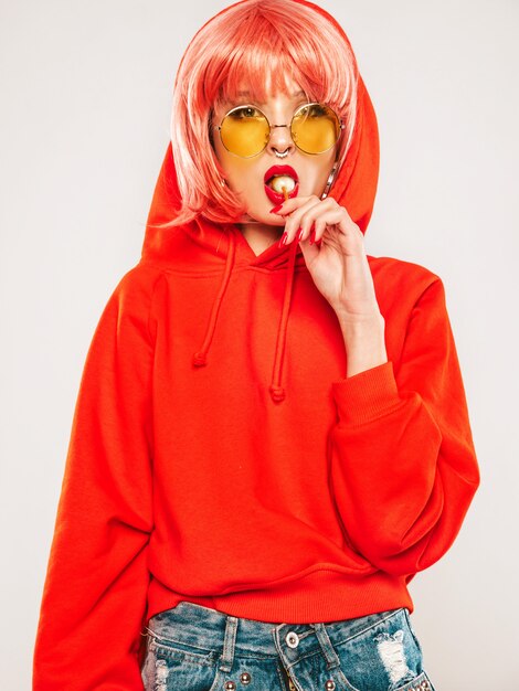 Retrato de joven hermosa hipster chica mala en moda verano rojo con capucha y arete en la nariz. Mujer rubia sonriente despreocupada sexy posando en el estudio con peluca. Modelo positivo lamiendo dulces de azúcar redondo